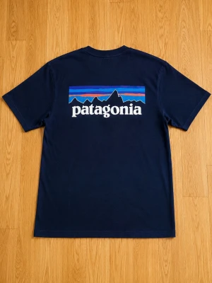 Mörkblå Patagonia t-shirt - Snygg mörkblå t-shirt från Patagonia med klassisk logga och bergmotiv i blått, lila och orange på ryggen samt liten logga fram. T-shirten har rund hals och korta ärmar, perfekt för en avslappnad stil. Tillverkad i mjuk bomull.