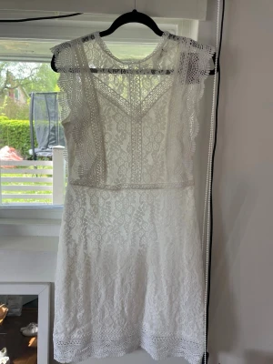 Vit studentklänning i spets med volangdetaljer - Superfin vit kortklänning i spets med vackra volangdetaljer längs axlarna och ärmlös design. Klänningen har hög hals och fina spetsmönster över hela plagget, perfekt för somriga dagar eller speciella tillfällen.
