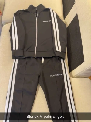 Svart loungewear-set Palm Angels - Snyggt svart loungewear-set från Palm Angels med vita ränder längs ärmar och ben. Setet består av en zip-jacka och matchande byxor, båda med Palm Angels-logga tryckt i vitt. Perfekt för en avslappnad och trendig stil.