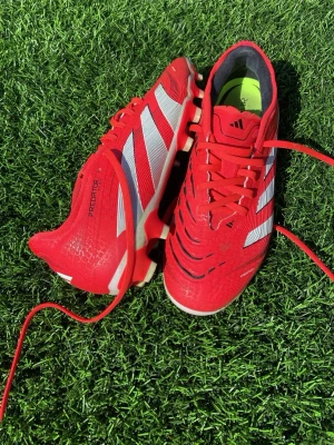 Adidas Predator röda fotbollsskor - Säljer ett par röda Adidas Predator fotbollsskor med vita och svarta detaljer. Skorna har snörning, klassisk låg profil och mönstrad ovandel för bättre bollkontroll. Snygg design med tydliga Adidas-ränder på sidorna. Bara använda ett par gånger. NY pris 1500