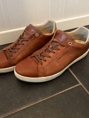 Bruna sneakers från Björn Borg - Snygga bruna sneakers från Björn Borg i äkta läder med klassisk låg profil och vita sulor. Skorna har bruna skosnören, diskret logga på sidan och en bekväm innersula. Perfekta för dig som gillar stilrena och tidlösa sneakers.