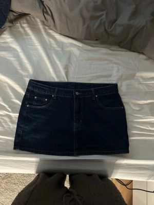 Mörkblå jeanskjol i klassisk modell - Snygg mörkblå jeanskjol med klassisk femficksdesign och hällor för bälte. Kjolen har en rak, kort passform och är tillverkad i denim. Perfekt för dig som gillar en tidlös och enkel stil. Den är lågmidjad och perfekt nu för sommaren. 