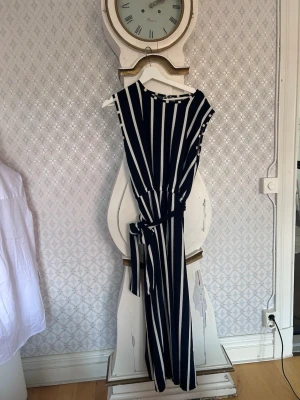 Randig blåvit jumpsuit med knytskärp - Snygg blåvit jumpsuit med vertikala ränder och knytskärp i midjan.  Perfekt för dig som vill ha en trendig och bekväm look. Materialet känns mjukt och följsamt.