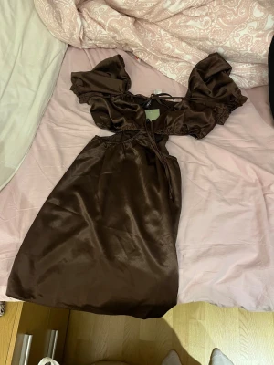 Brun satinklänning med puffärmar - Snygg brun klänning i satin med puffiga kortärmar och knytning framtill. Klänningen har en omlott-detalj vid bysten och en lätt glansig finish som ger en lyxig känsla. Perfekt för dig som vill sticka ut med en trendig look. helt ny aldrig använd. 