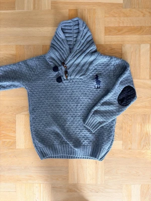 Grå stickad tröja från Ralph Lauren - Snygg grå stickad hoodie från Ralph Lauren med quiltade svarta armbågslappar och ribbade muddar. Tröjan har en hög krage med knappar och trädetaljer samt den klassiska broderade loggan på bröstet. Perfekt för en chill och stilren look. Priset är inte hugget i sten! Kom gärna med pris förslag! 😁🙌🤝