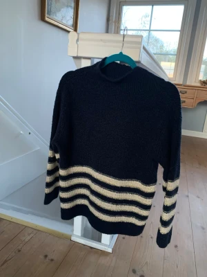 Svart stickad tröja med beige ränder - Mysig svart stickad tröja med breda beige ränder på nedre delen av kroppen och ärmarna. Tröjan har en hög hals och en avslappnad passform, perfekt för kyliga dagar. Klassisk design som funkar till många olika stilar.