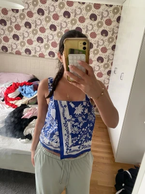 Blå blommig linnetopp - Säljer en blå linnetopp med vita blommor och breda axelband. Toppen har ett unikt omlottliknande snitt framtill och är croppad. Perfekt för dig som vill ha en somrig och trendig look.