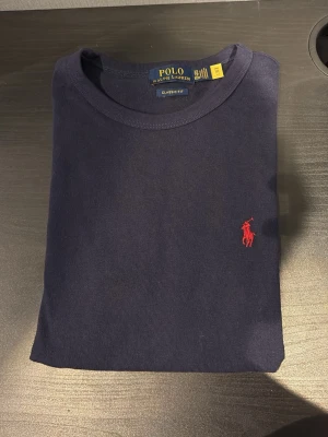 Mörkblå t-shirt från Polo Ralph Lauren - Snygg mörkblå t-shirt från Polo Ralph Lauren med klassisk passform och rund hals. T-shirten har det ikoniska röda broderade logotypen på bröstet och är tillverkad i mjuk bomull. Perfekt för en stilren och avslappnad look.