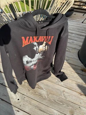 Svart Makaveli hoodie med tryck - Säljer en svart hoodie med stor huva och dragsko, prydd med ett stort Makaveli-tryck och bild på Tupac på framsidan. Hoodien har känguruficka och ribbade muddar. Perfekt för dig som gillar streetwear och hiphop-vibe.