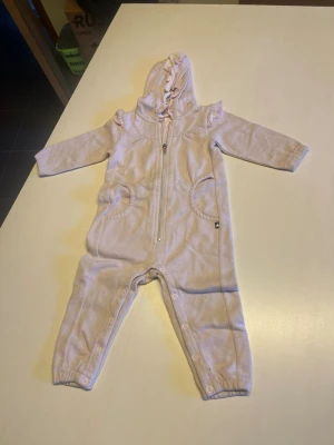 Ljusrosa byxdress med volang och huva - Supersöt ljusrosa byxdress med huva och volangdetaljer på axlarna. Dragkedja framtill och tryckknappar längs benen för enkel påklädning. Mjukt material och elastiska muddar vid ärmar och benslut. Perfekt för mysiga dagar.