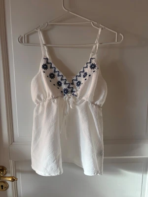 Vit broderad linnetopp med knyt - Söt vit linnetopp med tunna axelband och blå broderade blommor längs v-ringningen. Toppen har knyt i midjan och är tillverkad i ett lätt, somrigt material. Perfekt för en avslappnad och trendig look.