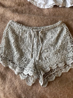 Vita virkade shorts med snörning - Superfina vita virkade shorts med snörning i midjan och vacker spetsdetalj. De har blommigt mönster och vågig kant nertill, perfekta för sommarens varma dagar. Lätta och luftiga, passar perfekt till en avslappnad stil.
