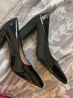 Svarta lackpumps med blockklack - Stilrena svarta pumps i glansigt lackmaterial med fyrkantig tå och bred blockklack. Perfekta för dig som vill ha en klassisk men trendig look. Skorna har en elegant siluett och ger en modern touch till outfiten.