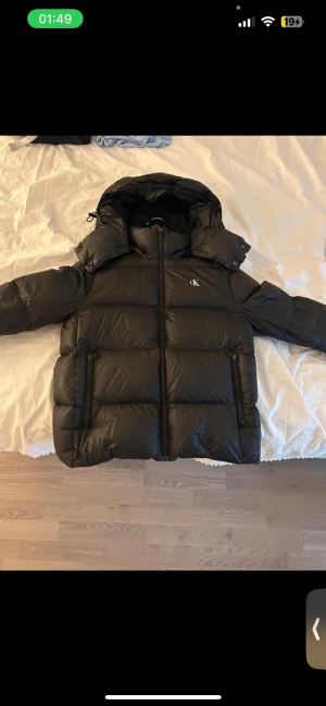 Svart pufferjacka från Calvin Klein - Säljer en svart pufferjacka från Calvin Klein med huva och dragkedja framtill. Jackan har två stora fickor med dragkedja och diskret CK-logga på bröstet. Perfekt för kalla dagar med sin vadderade design och klassiska look. Den har vid ett tillfälle slipats nere på vänster sida ( se bild ) 