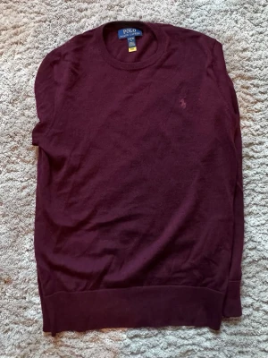 Vinröd stickad tröja Polo Ralph Lauren - Vinröd stickad tröja från Polo Ralph Lauren med rund halsringning och diskret broderad logga på bröstet. Tröjan har långa ärmar och ribbade muddar vid ärmslut och nederkant. Perfekt för lager på lager och klassisk stil.