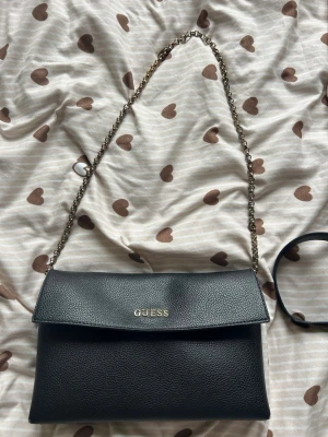 Svart axelväska från Guess - Snygg svart axelväska från Guess i slätt konstläder med guldfärgad kedjerem. Väskan har lock med magnetknäppning och ett innerfack med dragkedja. Perfekt storlek för det viktigaste och stilren design med Guess-logga framtill.