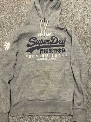 Grå hoodie från Superdry - Snygg grå hoodie från Superdry med stor logga och japansk text på bröstet. Klassisk känguruficka framtill, vita dragsnören i huvan och ribbade muddar. Perfekt för en avslappnad streetstyle-look.