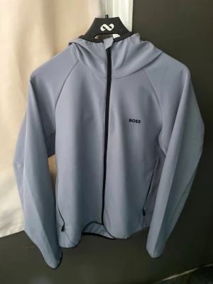 Ljusblå hoodie från BOSS - Snygg ljusblå hoodie från Hugo boss med dragkedja framtill och diskret logga på bröstet. Hoodien har långa ärmar, huva och svarta detaljer längs dragkedjan och muddar. Perfekt för en stilren och avslappnad look.