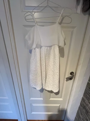 Vit kort klänning med volangdetalj - Söt vit kort klänning med smala axelband och volang upptill. Kjolen har en strukturerad, nästan broderad känsla och framtill finns en dekorativ brosch. Perfekt för sommarens alla tillfällen.