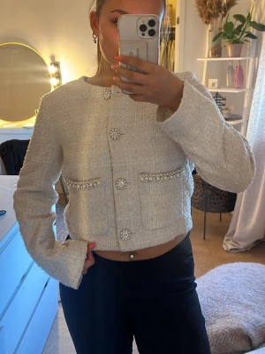 Vit tweedkavaj med pärldetaljer - Trendig vit kavaj i tweed med raka axlar och croppad passform. Kavajen har dekorativa pärlknappar och fickor med pärl- och stenbroderier framtill. Perfekt för en chic och stilren look. Köpt på Sellpy men är kommer ifrån hm 