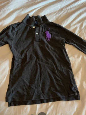 Svart/grå långärmad pikétröja Polo Ralph Lauren - Svart/grå långärmad pikétröja från Polo Ralph Lauren med klassisk krage och knappar framtill. Stor lila logga broderad på bröstet. Perfekt för en stilren och avslappnad look. Xs/s