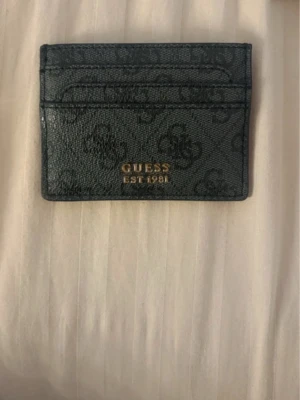  korthållare  Guess - Snygg svart korthållare från Guess med logomönster och guldfärgad text 'GUESS EST 1981' på framsidan. Tillverkad i konstläder med flera kortfack, perfekt för att hålla ordning på dina kort.