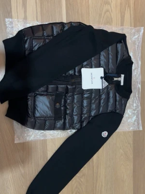 Moncler cardigan - Snygg svart pufferjacka från Moncler med quiltad front och två stora fickor med lock och knapp. Jackan har svarta ärmar i mjukt material och ribbade muddar. Klassisk Moncler-logga på ärmen och dragkedja framtill. Perfekt för kalla dagar.