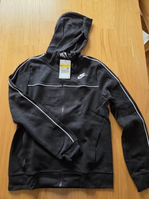 Nike svart dam huvtröja, storlek S - Svart huvtröja med dragkedja och vita detaljer längs ärmarna och över bröstet. Tröjan har en vit Nike-logotyp på vänster bröst och fickor framtill. Huvan är justerbar med dragsko.