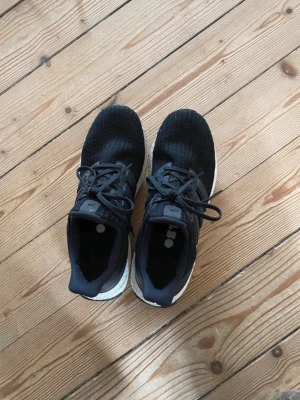 Adidas Ultra Boost svarta sneakers - Svarta Adidas Ultra Boost sneakers med stickad ovandel och vita Boost-sulor. Klassisk design med tre svarta ränder på sidan och snörning. Sköna och lätta skor som passar perfekt till streetwear och träning.