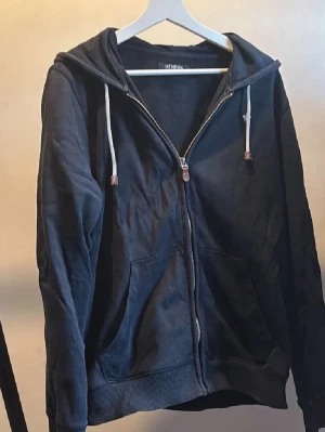 Morris Mörkblå zip-hoodie  - Mörkblå hoodie från Morris med dragkedja framtill, två stora fickor och huva med vita dragsnören och bruna detaljer. Tillverkad i mjukt material och har ribbade muddar vid ärmslut och nederkant. Perfekt för en avslappnad stil.