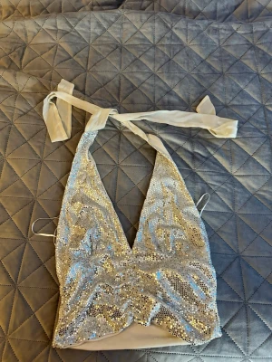 Glittrig silver topp med knytband - Snygg glittrig topp i silver från GinaTricot med paljetter och djup v-ringning. Toppen har knytband i nacken och är ärmlös, perfekt för att sticka ut. Passar dig som gillar att glänsa och vill ha en trendig look.