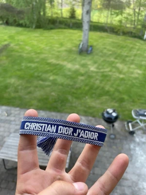 Blått Christian Dior J'Adior armband - Snyggt blått tygarmband från Christian Dior med texten 'J'Adior' broderad i vitt. Armbandet har justerbar knytning och dekorativa tofsar i ändarna. Perfekt accessoar för att lyfta din outfit med en lyxig touch.