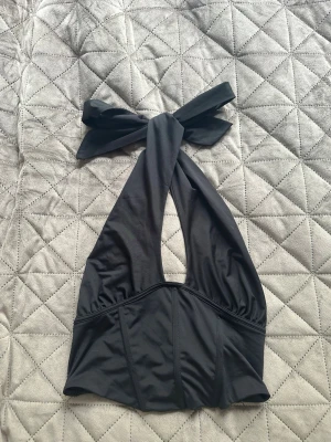 Svart halterneck från Gina Tricot - Svart halterneck med corsett likande midja från Gina Tricot med breda band som knyts i nacken. Modellen är croppad och har en stilren, ärmlös design i mjuk polyester. Perfekt för en utgång.