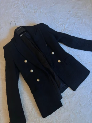 Svart kavaj med guldknappar från ZARA - Stilren svart kavaj från ZARA med sex dekorativa guldknappar framtill. Kavajen har markerade axlar, klassisk krage och två fickor. Materialet är strukturerat och ger en lyxig känsla. Perfekt för en trendig och uppklädd look.