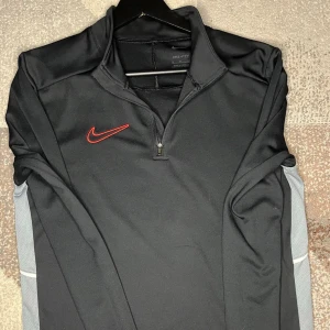 nike träningströja - Svart långärmad träningströja från Nike med halv dragkedja framtill och röd Nike-logga på bröstet. Grå paneler längs sidorna ger en sportig look. Perfekt för träning eller uppvärmning. Tillverkad i ett mjukt och stretchigt funktionsmaterial.