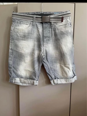 Grå jeansshorts från Jack&jones S nya  - Snygga grå jeansshorts från Louis Vuitton med slitna detaljer och uppvikta benslut. Shortsen har fem fickor och ett rutigt bälte med metallspänne där Louis Vuitton-loggan syns. Perfekta för en avslappnad och trendig look.
