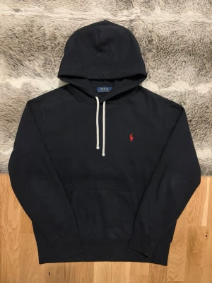 Svart Polo Ralph Lauren Fleece Hoodie - Storlek M - Hej! 👋 Säljer en klassisk svart Polo Ralph Lauren hoodie i storlek M med röd pony-logga. 🐎 Tröjan är i mycket bra skick i övrigt, men har ett par mindre hål/slitningar i tyget på huvan (se de två sista bilderna). Priset är justerat efter detta! 100% äkta med originaltvättlappar kvar och en fullt fungerande QR-kod för enkel verifiering ✅ Perfekt, prisvärt premium-basplagg! Fraktar snabbt! 🚛📦💬 