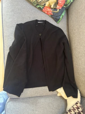 Svart kofta från Gina Tricot - En klassisk svart kofta från Gina Tricot med rund halsringning och en knapp framtill. Koftan har långa ärmar och ribbade kanter. Perfekt att slänga över en t-shirt eller klänning för en enkel och stilren look.
