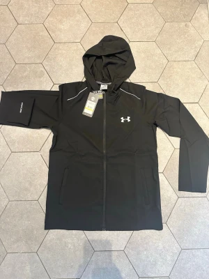 Under Armour Vanish Performance Jacket - Snygg svart vindjacka från Under Armour med huva och hel dragkedja. Jackan har vita detaljer, logga på bröstet och stort UNDER ARMOUR-tryck på ryggen. Tillverkad i vattenavvisande material med två fickor framtill och elastiska muddar. Perfekt för blåsiga dagar.