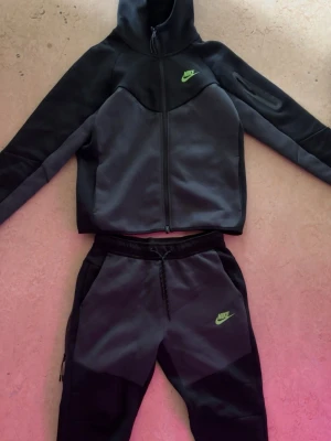 Nike Tech Tracksuit - Säljer ett par mjukisbyxor från Nike i svart och mörkgrå färg med neongrönt Nike-logo på vänster ben. Byxorna har dragsko i midjan, sidofickor och är tillverkade i ett mjukt och bekvämt material som passar perfekt för chill eller träning. Snyggt snitt och modern design.