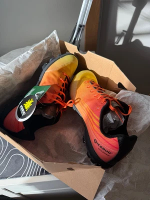 Icebug löparskor orange/gul - Snygga löparskor från Icebug i en cool färgskala som går från gult till orange med svarta detaljer. Skorna har grov sula för bra grepp, snörning och är tillverkade i slitstarkt syntetmaterial. Perfekta för terränglöpning och utomhusaktiviteter. Passar även storlek 42. Värde: 1499kr. Mitt pris: 499