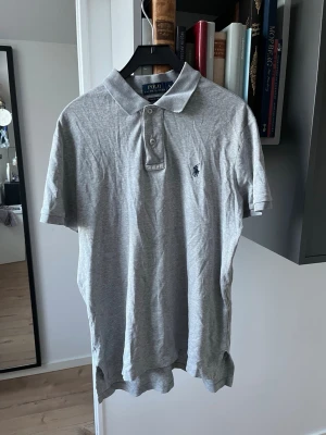 Grå pikétröja från Polo Ralph Lauren - Klassisk grå pikétröja från Polo Ralph Lauren med korta ärmar och krage. Tröjan har knappar framtill och den ikoniska broderade loggan på bröstet. Perfekt till jeans eller shorts för en avslappnad stil. Längre där bak än fram till. 