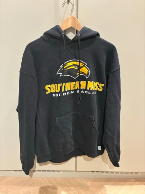 Svart Southern Miss hoodie - Svart hoodie med huva från Russell Athletic. Framsidan har tryck med Southern Miss Golden Eagles och en gul örn. Klassisk college-stil med dragsko i huvan och ribbade muddar. Perfekt för dig som gillar sportig och avslappnad look.