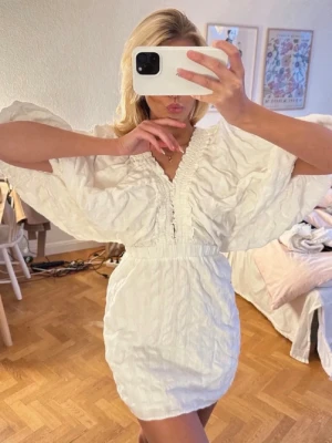 Vit kort klänning med volangärmar - Superfin vit klänning med kort längd och breda volangärmar. Djup v-ringning med rynkade detaljer framtill och markerad midja. Perfekt för en trendig och fräsch look.