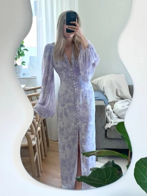 Adoore Postiano dress - Superfin ljuslila maxiklänning från Adoore med vackert mönster, långa puffärmar och djup v-ringning. Klänningen har knappar framtill och en slits som ger en snygg detalj.  Sista bilden är precis under dragkedjan på baksidan