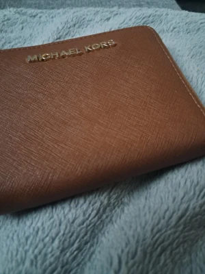 Brun plånbok från Michael Kors - Snygg brun plånbok från Michael Kors i ett stilrent och klassiskt utförande. Plånboken är gjord i strukturerat läder med guldfärgad logotyp på framsidan. Perfekt för att hålla ordning på kort och kontanter.
