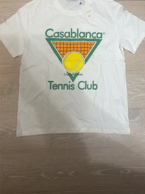 Vit Casablanca Tennis Club t-shirt - Vit t-shirt från Casablanca med stort färgglatt tryck framtill som visar en tennisboll och texten 'Tennis Club'. Klassisk passform med rund hals och korta ärmar. Perfekt för dig som gillar sportig stil och grafiska prints.