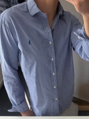Blårutig skjorta från Polo Ralph Lauren - Klassisk blårutig skjorta från Polo Ralph Lauren med lång ärm och knappar framtill. Skjortan har en liten broderad logga på bröstet och är tillverkad i mjuk bomull. Perfekt för en stilren och avslappnad look. Storlek 39 vilket motsvarar m