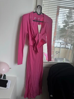 Rosa ribbad långklänning från adoore  - Trendig rosa långklänning från Adoore med ribbad struktur och lång ärm. Klänningen har en snygg knytning framtill och en klassisk krage. Perfekt för dig som vill sticka ut med en färgstark och modern look.