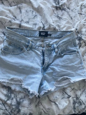Ljusblå denimshorts från 157 - Säljer ett par ljusblå denimshorts från 157 med fransiga benslut och slitna detaljer framtill. Klassisk femficksmodell med knapp och dragkedja. Perfekta för varma dagar och enkel att matcha med olika toppar.